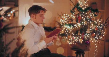 Yakışıklı genç adam, arka planda ışıklandırılmış Noel ağacıyla evdeki sandalyede otururken kitap okuyor.