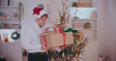 Eve çeşitli Noel hediyeleri taşıyan resmi ve Noel Baba şapkası takan yakışıklı bir genç adam portresi.