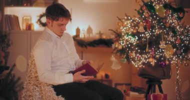 Genç adam, arka planda ışıklandırılmış Noel ağacıyla evdeki sandalyede otururken kitap okuyor.