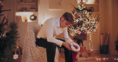Noel Baba 'nın şapkasını tutan üzgün genç adam arka planda ışıklandırılmış Noel ağacıyla evde otururken