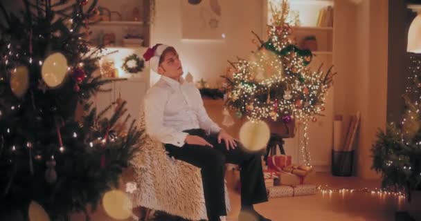 クリスマスの間に照らされた家の椅子に座っている間サンタの帽子を取り除く形態の心配な若者 — ストック動画