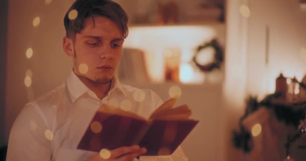 ハンサムな若者はクリスマスの間に家に座って本を読む — ストック動画