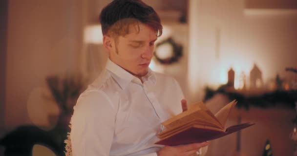Guapo Joven Leyendo Libro Mientras Está Sentado Casa Durante Navidad — Vídeos de Stock
