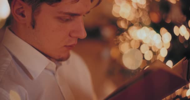 Pemuda Tampan Membaca Buku Sambil Duduk Rumah Diterangi Selama Natal — Stok Video