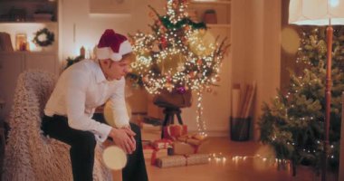 Resmi kıyafetler içindeki üzgün genç adam ve Noel Baba şapkası Noel boyunca aydınlatılmış evdeki sandalyede oturuyor.