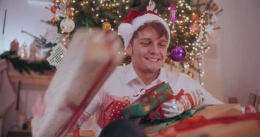 Noel Baba şapkalı heyecanlı genç adam süslenmiş evde çeşitli Noel hediyeleri topluyor.