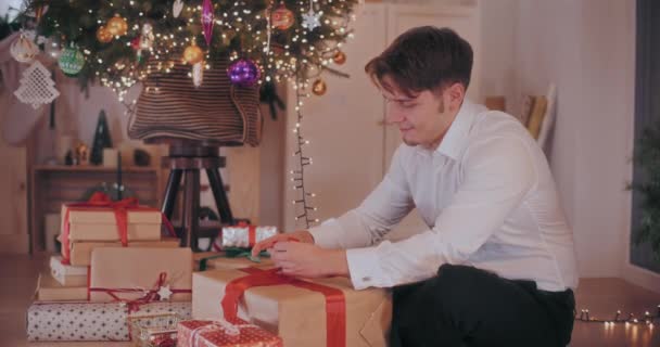 クリスマスの間に自宅で床に座っている間包まれたギフト用の箱にリボンを結ぶ形をした幸せな若者 — ストック動画