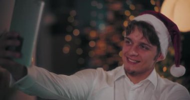 Noel Baba şapkalı genç adam Noel boyunca evde dijital tabletle selfie çekiyor.