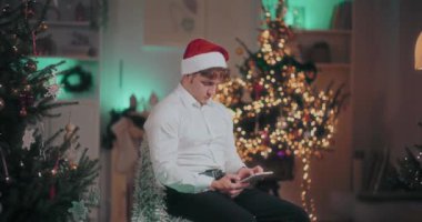 Noel Baba şapkalı genç adam, Noel 'de aydınlatılmış evdeki sandalyede otururken dijital tablet kullanıyor.