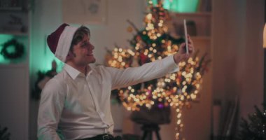 Mutlu genç adam tablet bilgisayarda selfie çekerken arka planda ışıklandırılmış Noel ağacı olan bir sandalyede oturuyordu.