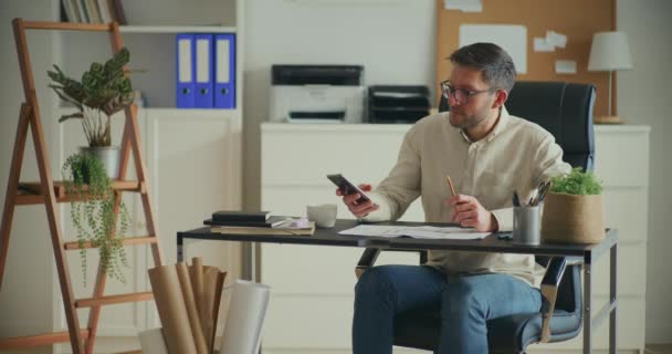 Młody Przedsiębiorca Sprawdza Smartfon Przygotowuje Dokumenty Siedząc Biurze — Wideo stockowe