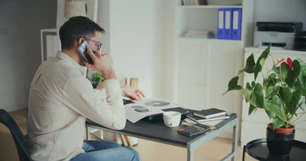 男性商业专业人员坐在办公桌前检查饼图和用智能手机交谈的侧视图 — 图库视频影像