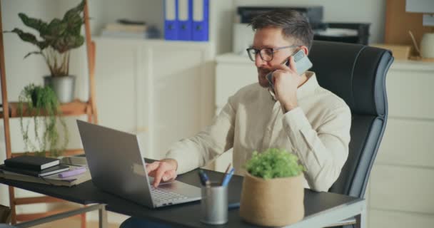 オフィスの机に座っている間 ノートパソコンを使用し 電話で話すビジネスマン — ストック動画