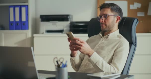 男性企业家坐在办公室的椅子上 一边用智能手机聊天 — 图库视频影像