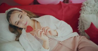 Galentines gününde kanepeye uzanmış kalp şeklinde şekerler tutan mutlu genç bir kadın.