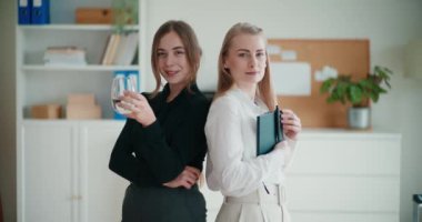 İş yerinde sırt sırta duran gülümseyen genç bayan meslektaşların portresi.