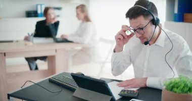 Kurumsal çalışma alanında hesap yaparken ve kağıda yazarken kulaklıkla konuşan kendine güvenen işadamı