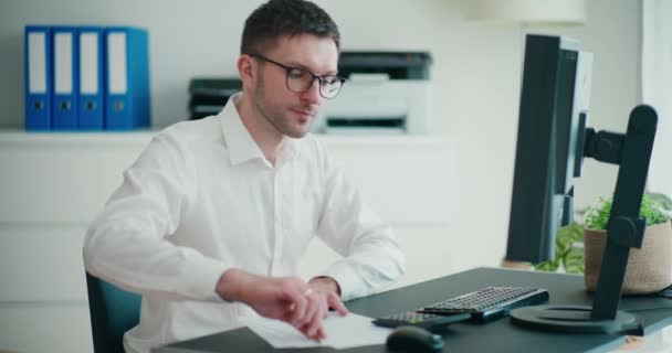若いビジネスマンは 紙に書いたり オフィスの机でコンピュータを使用しながら 予算を計算しています — ストック動画