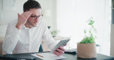 Stresli iş adamı, kurumsal çalışma alanındaki masasında oturan gözlükleri çıkarırken dijital tablet kullanıyor.
