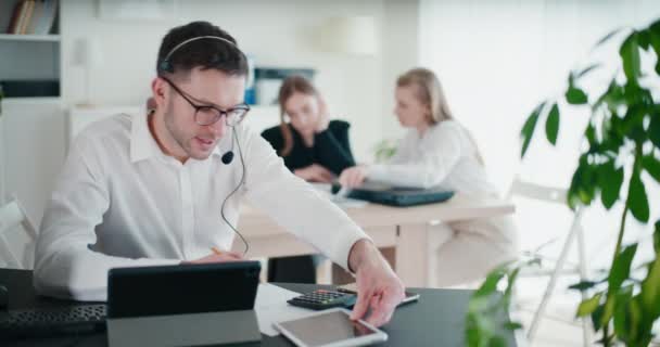 stock video Confident male entrepreneur talking through headset while calculating and writing on paper in corporate workspace