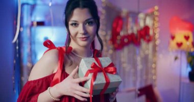 Galentines Günü 'nde aydınlatılmış bir evde elinde hediye paketi olan gülümseyen genç bir kadının portresi.