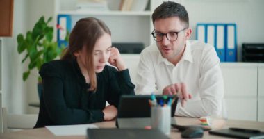 Genç işadamı, dijital tablet üzerinden stratejiyi açıklarken, işkadını ofisteki masada kağıda yazı yazıyor.