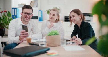 Genç iş adamı, şirket ofisindeki masada otururken gülümseyen bayan meslektaşlarıyla akıllı telefonu paylaşıyor.