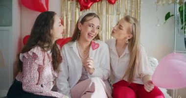 Evde Galentines günü kutlamalarında kalp şeklinde şekerler tutan mutlu genç kadınlar öpüşüyor.