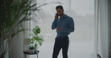 Kendine güvenen genç girişimci akıllı telefondan stratejiyi tartışırken çalışma alanının penceresinin önünde duruyor