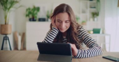 Evdeki oturma odasında masaya yaslanırken dijital tablet kullanan gülümseyen genç bir kadının kilit görüntüsü.