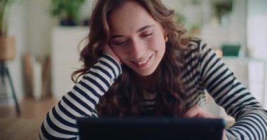Evdeki oturma odasında yatarken dijital tablet kullanan gülümseyen genç bir kadının kilit altında çekilmiş görüntüleri.