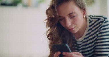 Oturma odasında uzanmış cep telefonuyla mesajlaşan genç bir kadın.