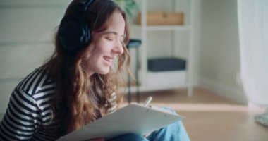 Evdeki oturma odasında kulaklıkla müzik dinlerken panoya resim çizen mutlu genç bir kadın.