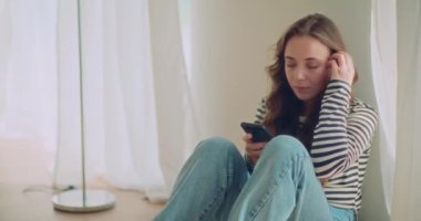 Evde oturma odasında otururken cep telefonuyla mesajlaşan genç bir kadın.