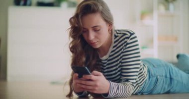 Genç bayan esmer, evde oturma odasında yerde yatarken akıllı telefon kullanarak mesaj atıyor.