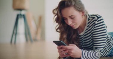 Evdeki oturma odasında yerde yatarken cep telefonundan mesaj atan mutlu genç kadın.