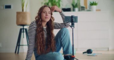Güzel, genç bir bayan içerik yaratıcısı kendini kameraya çekiyor. Evde akıllı telefonun önünde tripodla konuşuyor.