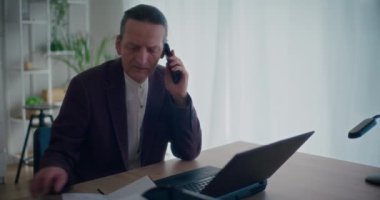 Üst düzey yönetici ofiste dizüstü bilgisayarda çalışırken cep telefonuyla konuşuyor ve not defterine yazıyor.