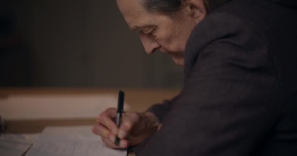 年配の男性マネージャーがオフィスで深夜勤務中にデスクで日記を書いている — ストック動画