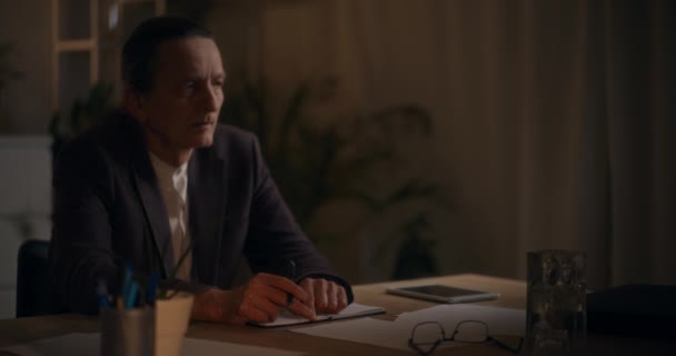 オフィスで深夜働いている間に机に日記をつけて座っている緊張した上級男性マネージャー — ストック動画