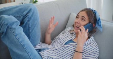 Neşeli bir kadın oturma odasındaki kanepeye uzanıp sohbet ederken cep telefonunda mutlu bir şekilde sohbet ediyor. Sohbet sırasında sıcaklık ve mutluluk saçıyor.