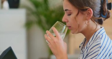 Çoklu görev yapan bir iş kadını özenle dizüstü bilgisayarı üzerinde çalışırken bardaktan su yudumluyor.