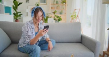 Neşeli ve memnun bir kadın, oturma odasının rahat ortamında kanepede dinlenirken akıllı telefonuna göz atmaktan zevk alırken gülümser.