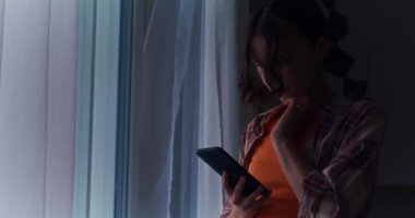 Sosyal medyaya kendini kaptırmış genç bir kız pencerenin kenarında duruyor, akıllı telefonunu ve interneti karıştırıyor, dijital ilişkinin etkisini gösteriyor.