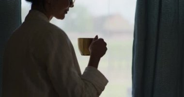 Pencerenin kenarında kahve içen, dinlenen ve anın tadını çıkaran bir kadının silüeti..