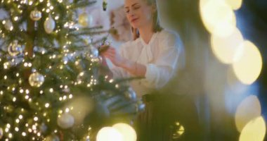 Gülümseyen ve zarif bir kadın dikkatlice süslenmiş ağacına bir Noel mücevheri yerleştirir..