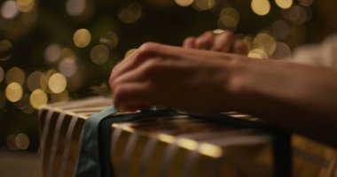 Noel hediyesini sarıp sarmalayan ellerin yakın çekimi. Arkaplanda parlayan bir ağaç ve ışıklar var..