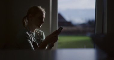 Mutfak penceresinin yanında telefonunu karıştıran bir kadının ana hatları..