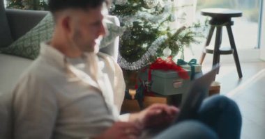 Ev bilgisayarında çalışan bir iş adamının silueti, Noel hediyelerinin arkaplanına karşı çerçevelenmiş ve süslenmiş bir Noel ağacı, şenlikli bir çalışma alanı yaratıyor..