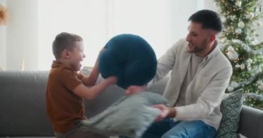 Bir baba ve oğlu, Noel 'de kanepenin üzerinde eğlenceli bir yastık savaşı yapıp neşeli, neşeli ve sıcak aile anlarını neşeli bir atmosferde paylaşıyorlar..